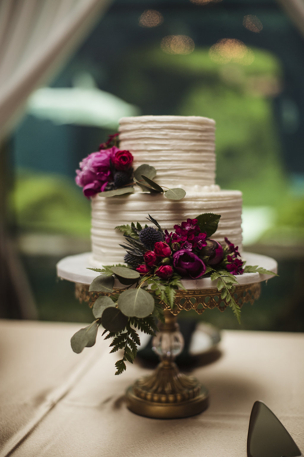 Two Tier Round White Wedding Cake on Ornate Gold Cake Stand with Fuchsia and Red Flowers with Greenery