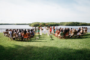 Outdoor Waterfront Wedding Ceremony Portrait with Wooden Folding Chairs, Hurricane Lanterns, and Tropical Floral and Greenery Ceremony Arch, Bridesmaids in Clover Green and Guava Orange Dresses | Tampa Bay Hotel Wedding Venue DoubleTree Suites by Hilton