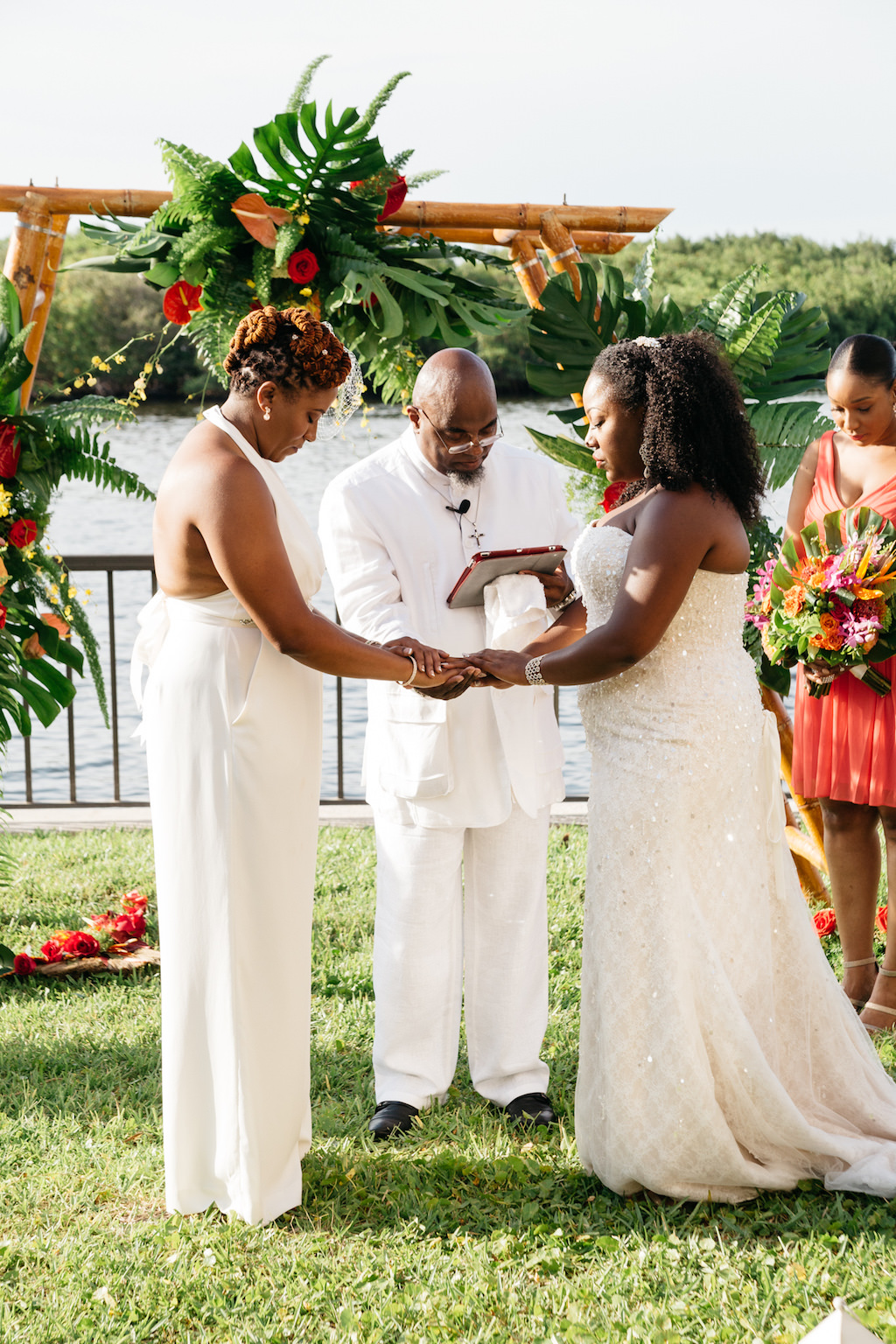 Outdoor Waterfront Wedding Ceremony Portrait, Brides in BHDLN Halter Pantsuit and Strapless Davids Bridal Dress, with Bamboo Ceremony Arch with Red and Orange Florals with Tropical Greenery