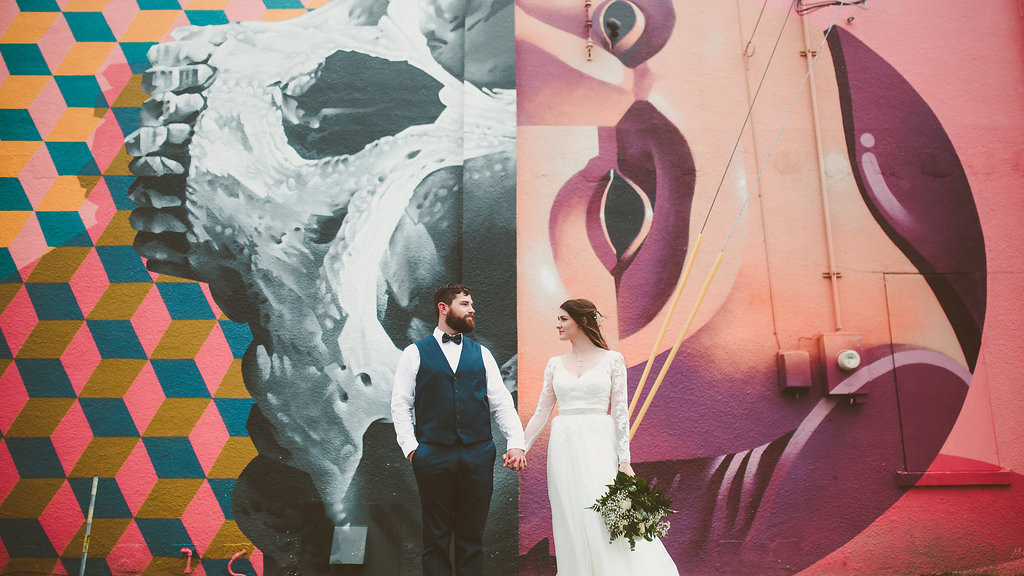 Outdoor Industrial Bridal Portrait in Lace Long Sleeve Belted Davids Bridal Wedding Dress with Green Fern and White Floral Bouquet with Colorful Street Graffiti Mural Art | Downtown St Pete