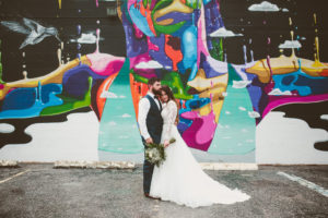 Outdoor Industrial Bridal Portrait in Lace Long Sleeve Belted Davids Bridal Wedding Dress with Green Fern and White Floral Bouquet with Colorful Street Graffiti Mural Art | Downtown St Pete