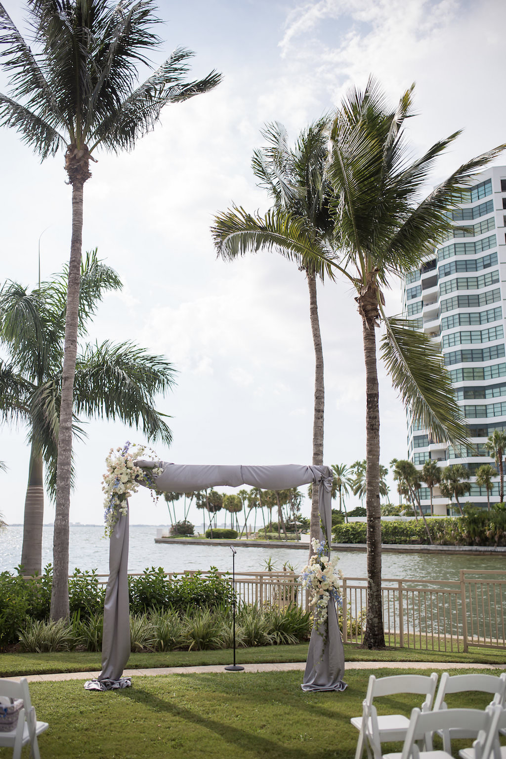 Outdoor Waterfront Downtown Hotel Wedding Ceremony with White Folding Chairs, Gray Silk Draped Ceremony Arch with White Florals | Sarasota Wedding Venue The Ritz Carlton