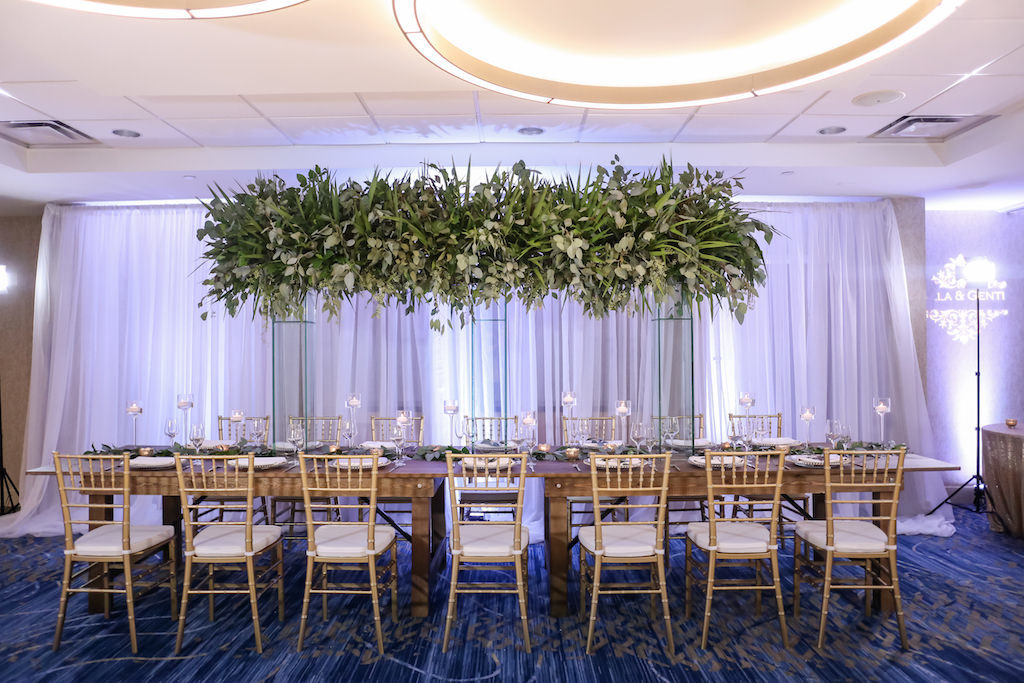 Hotel Ballroom Wedding Reception With Long Wood Feasting Table, Extra Tall Greenery Centerpiece in Clear Glass Rectangular Vases, Gold Chiavari Chairs, White Draping, and Glass Candleholders at Varying Heights | Beach Hotel Wedding Venue Hilton Clearwater Beach