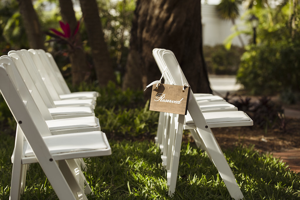 Outdoor Tropical Garden Wedding Ceremony with White Folding Chairs, White Script on Wooden Sign hanging from Ribbon | Venue Longboat Island Chapel
