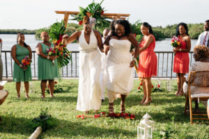 Outdoor Waterfront Wedding Ceremony Portrait, Brides in Halter BHDLN Pantsuit and Beaded Strapless Davids Bridal Dress, with Wooden Folding Chairs, Hurricane Lanterns, and Tropical Floral and Greenery Ceremony Arch, Bridesmaids in Clover Green and Guava Orange Dresses | Tampa Bay Hotel Wedding Venue DoubleTree Suites by Hilton