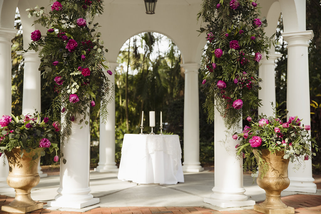 Outdoor Tropical Garden Gazebo Wedding Ceremony with Fuchsia and Pink Floral with tropical Greenery, Large Gold Classical Planters, and Pillar Candles | Venue Longboat Island Chapel | Sarasota Wedding Planner Jennifer Matteo Event Planning