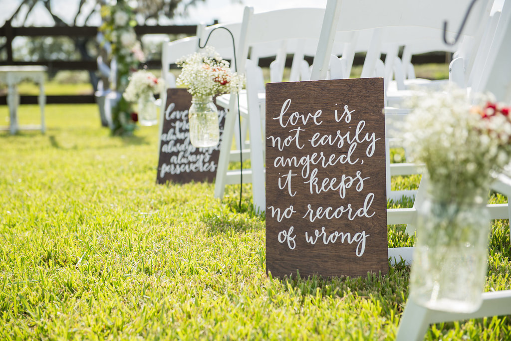 Outdoor Rustic Chic Wedding Ceremony with Love QUotes in Handpainted in White on Wooden Board, with Babys Breath and REd Floral in Hanging Mason Jars and White Folding Chairs