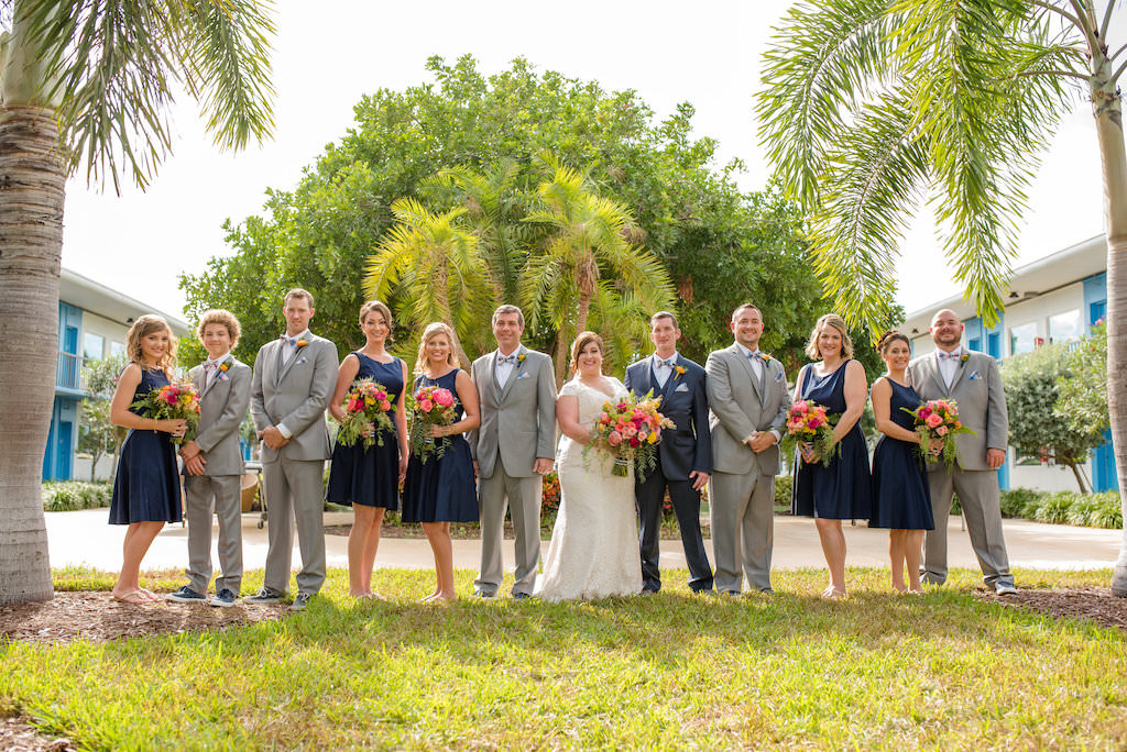 Outdoor Wedding Party Portrait, Bridesmaids in Tea Length Navy Blue Zazzle Dresses, Groomsmen in Gray Suits with Orange Boutonniere, Bride in V Neck Cap Sleeve Lace Dress from Truly Forever Bridal, with Tropical Pink Floral and Greener Bouquet | Tampa Bay Wedding Photographer Caroline and Evan Photographer | St Pete Beach Venue The Postcard Inn