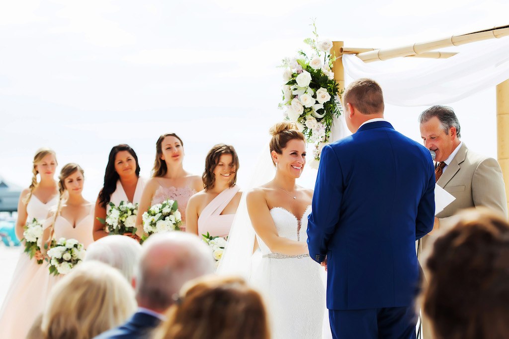 Outdoor Beach Wedding Ceremony, Bridesmaids in Mismatched Blush Dresses, Groom in Blue Suit, Bride in Strapless Belted Dress with Bamboo Ceremony Arch with WHite Flowers and Natural Greenery and White Draping