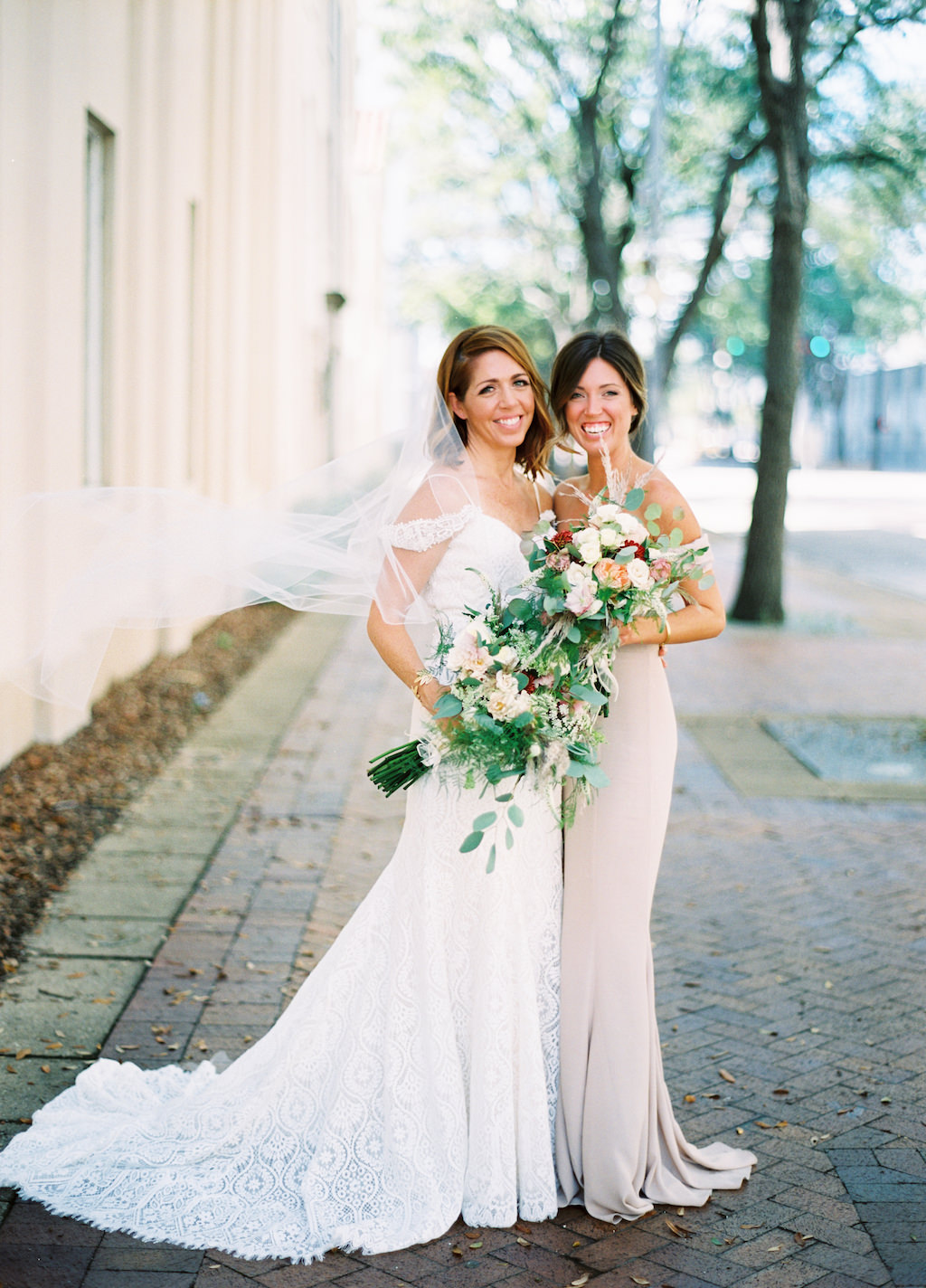 Outdoor Downtown Bride and Bridesmaid Portrait with Blush Pink, Red, and White Floral with Natural Greenery Bouquet, Bridesmaid in Ivory Column Revolve Dress, Bride in Off The Shoulder Madeline Gardner Dress