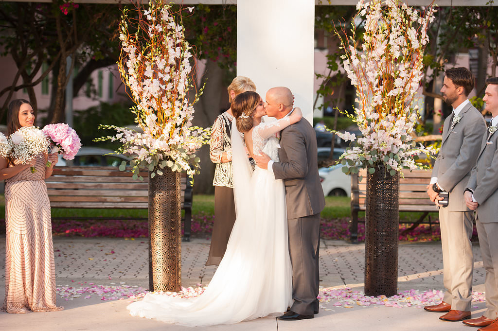 Outdoor Wedding Ceremony First Kiss Portrait, Groomsmen in Light Gray Suits, Bridesmaids in Blush Beaded Vintage Inspired Adrianna Papell Dress with Baby's Breath and Pink Peony Bouquet, Tall White Floral Arrangements with White Blossoms and Branches in Patterned Metal Planters, and Rose Petals | Downtown St Pete Venue North Straub Park