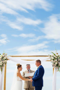 Outdoor Beach Wedding Ceremony, Groom in Blue Suit, Bride in Strapless Belted Dress with Bamboo Ceremony Arch with WHite Flowers and Natural Greenery and White Draping