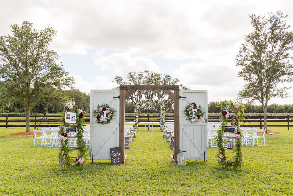 Outdoor Rustic Chic Wedding Ceremony Decor with Freestanding White Barn Door Gate with Oversized Initial Letters and Floral Wreath, Red, Pink and White Florals with Greenery Garland, and White Folding Chairs | Tampa Bay Farm Wedding Venue Wishing Well Barn