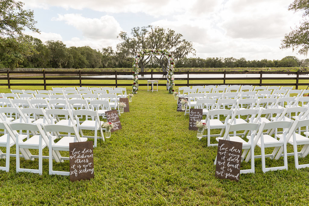 Outdoor Rustic Chic Wedding Ceremony with Love Quotes Hand Painted in White on Wooden Board, with Babys Breath and REd Floral in Hanging Mason Jars and White Folding Chairs, and Floral Ceremony Arch | Tampa Bay and Hillsborough County Farm Wedding Venue Wishing Well Barn
