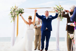 Outdoor Beach Wedding Ceremony, Groom in Blue Suit, Bride in Strapless Belted Dress with Bamboo Ceremony Arch with White Flowers and Natural Greenery and White Draping | Hotel Wedding Venue Hilton Clearwater Beach
