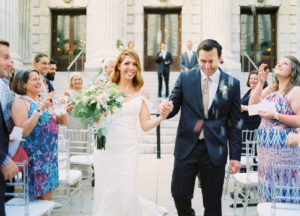 Outdoor Architectural Wedding Ceremony Exit Portrait on Front Steps of Downtown Tampa Hotel Venue Le Meridien, Bride in Madeline Gardner Dress with White FLoral and Natural Greenery Bouquet, Groom in Navy Suit with Champagne Tie and White and Greenery Boutonniere