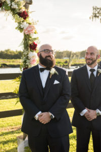 Outdoor Wedding Ceremony Portrait in front of Fence and Red, Whtie, and Pink Floral with Ferns and Greenery and Baby's Breath Arch, Groom in Black Suit with Boutonniere and Bowtie | Tampa Bay Rustic Wedding Venue Wishing Well Barn