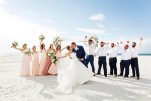Outdoor Beach Wedding Party Portrait, Bridesmaids in Mismatched Blush Dresses, Groom in Blue Suit, Bride in Strapless Belted Dress with Bamboo Ceremony Arch with WHite Flowers and Natural Greenery and White Draping | Tampa Bay Waterfront Hotel WEdding Venue Hilton Clearwater Beach