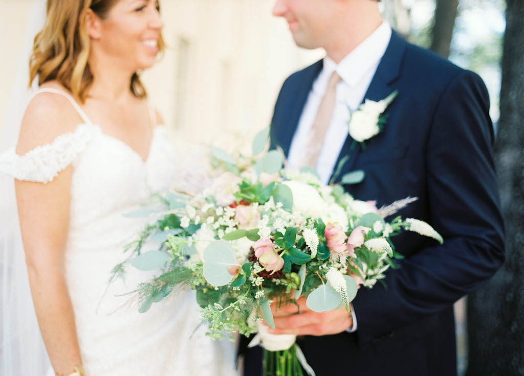 Outdoor Wedding Portrait, Bride in Off the Shoulder Madeline Gardner Dress, Groom in Navy Suit with Champagne Tie and White Boutonniere with Greenery, with White, Pink, and Red Floral with Natural Greenery Bouquet