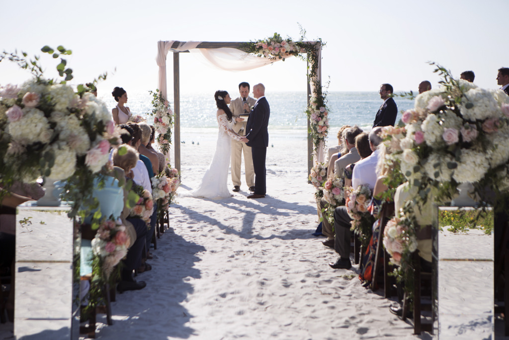 Siesta Beach Wedding Ceremony Portrait with Wood Folding Chairs, Peach, BLush, and White Rose with Natural Greenery Florals on Mirror Pedestals, and Ceremony Arch with Pink Draping, Bride in Lace Applique Long Sleeve Dress | Sarasota Wedding Photographer Djamel Photography