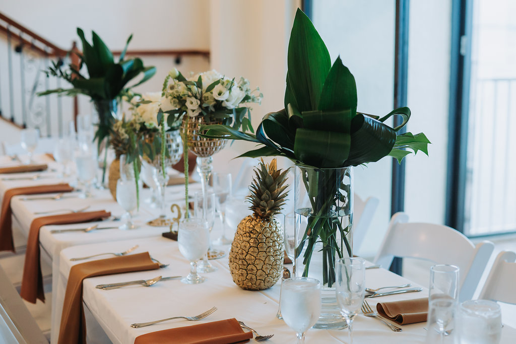Indoor Hotel Ballroom Wedding Reception with Tan and White Table Linens, Pineapple and Tall Tropical Greenery and White Floral Centerpieces in Glass Cylinder and Mirrored Vases, and White Folding Chairs | Tampa Bay Waterfront Venue Beso Del Sol