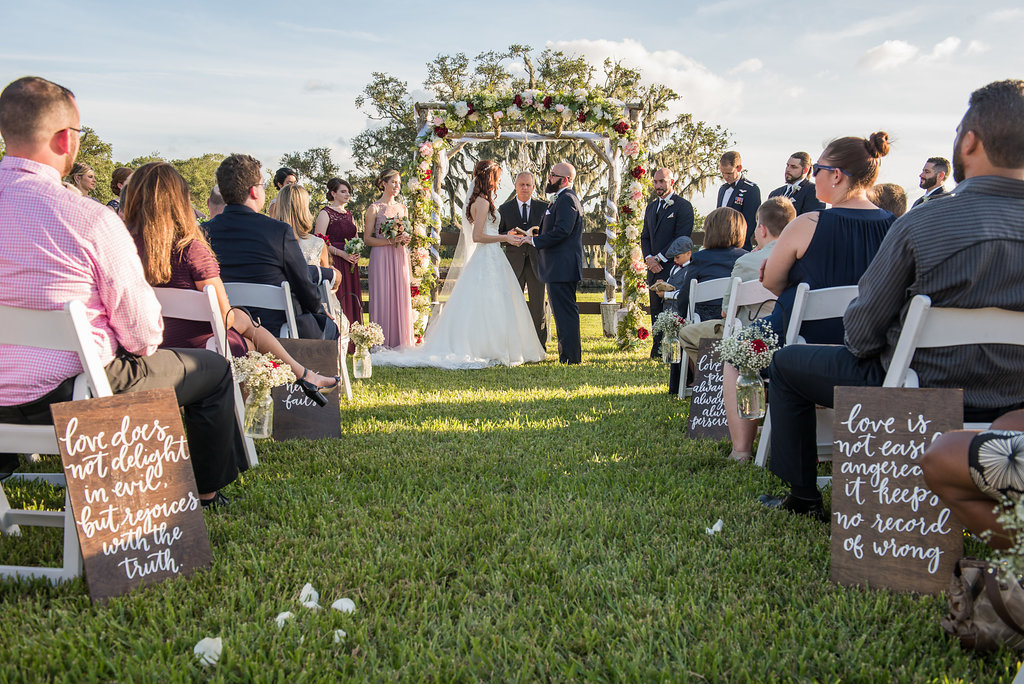 Outdoor Rustic Chic Wedding Ceremony with Love Quotes Hand Painted in White on Wooden Board, with Baby's Breath and Red Florals in Hanging Mason Jars and White Folding Chairs, and Floral Ceremony Arch, Bride in David's Bridal Ballgown Dress | Tampa Bay and Hillsborough County Farm Wedding Venue Wishing Well Barn