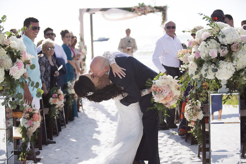 Siesta Beach Wedding Ceremony First Kiss Portrait with Wood Folding Chairs, Peach, BLush, and White Rose with Natural Greenery Florals on Mirror Pedestals, and Ceremony Arch with Pink Draping, Bride in Lace Applique Long Sleeve Dress | Sarasota Wedding Photographer Djamel Photography