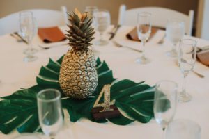 Tropical Wedding Reception with Brown and White Table Linens, Gold Painted Pineapple Centerpiece on Palm Leaf Greenery, and Gold Glitter Table Number and White Folding Chairs