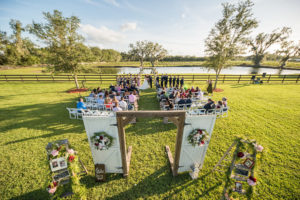 Outdoor Rustic Chic Wedding Ceremony Portrait with Freestanding White Barn Door Gate with Oversized Initial Letters and Floral Wreath, Red, Pink and White Florals with Greenery Garland, and White Folding Chairs | Tampa Bay Farm Wedding Venue Wishing Well Barn