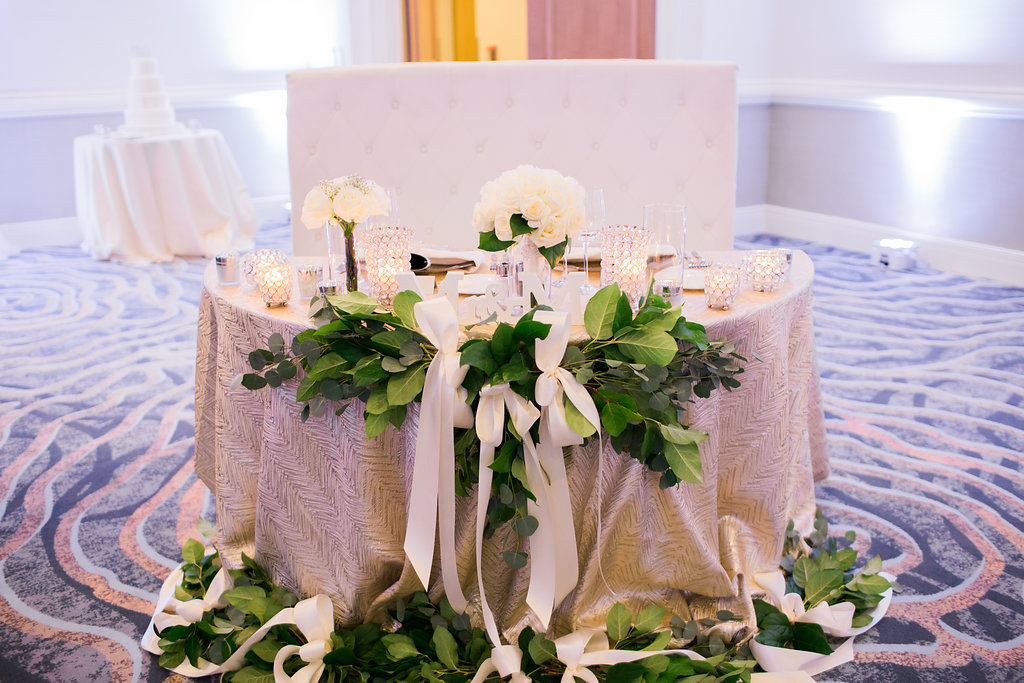 Elegant Wedding Reception Sweetheart Table with Textured Gold Linen, Greenery With White Ribbon Bows, Small White Rose Centerpiece and Cut Crystal Candleholders and High-back White Loveseat | Tampa Bay Wedding Planner Special Moments Event Planning | Venue Hyatt Regency Clearwater Beach | LInens Over The Top Linen Rentals | Rentals Gabro Event Services | Florist Apple Blossoms Floral Designs