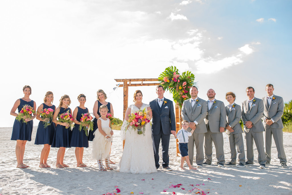 Outdoor Wedding Party Portrait, Bridesmaids in Tea Length Navy Blue Zazzle Dresses, Groomsmen in Gray Suits with Orange Boutonniere, Bride in V Neck Cap Sleeve Lace Dress from Truly Forever Bridal, with Tropical Pink Floral and Greenery Bouquet, Ring Bearer in Suspenders and Flower Girl in White Dress, and Bamboo Ceremony Arch | Tampa Bay Wedding Photographer Caroline and Evan Photographer | St Pete Beach Venue The Postcard Inn