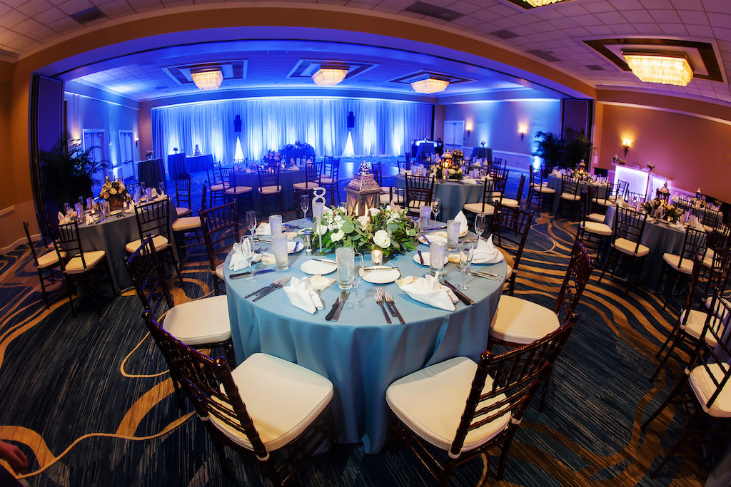 Indoor Hotel Ballroom Wedding Reception with Blue Table Linens, Low White Floral and Greenery Centerpiece with Hurricane Lantern, and Brown Chiavari Chairs | Waterfront Tampa Bay Hotel Wedding Venue Hilton Clearwater Beach | Rentals A Chair Affair
