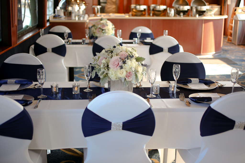 Indoor Cruise Wedding Reception with White OVal Backed Chairs with Navy Blue Wraps, Low Blush Pink and White ROse with Blue Berry and Greenery Centerpiece in Wooden Box, Blue Satin Table Runners, and Silver Chargers | Tampa Bay Unique Waterfront Wedding Venue Yacht Starship