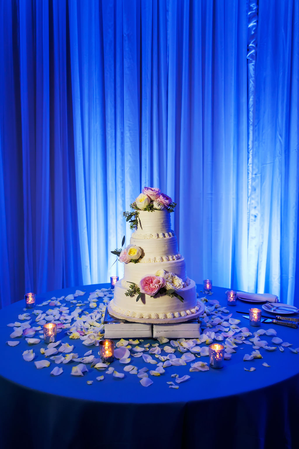 Four Tier Round White Wedding Cake on Book Cakestand with Pink and Yellow ROse with Greenery, Rose PEtals and Votive Candles on Blue Linen Table with Blue Draping and Uplighting