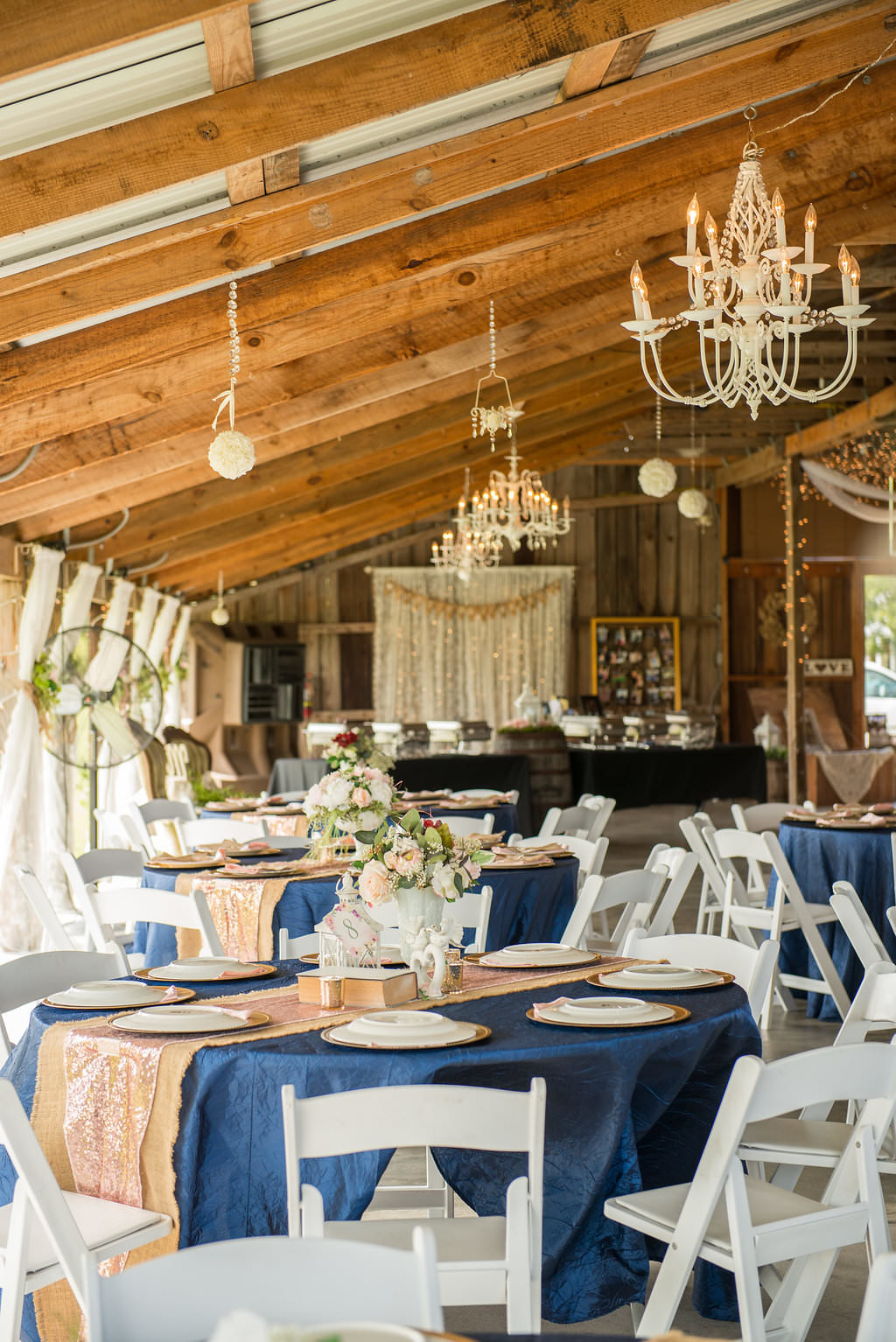 Rustic Wedding Reception with Round Tables with Navy Blue Linens and Burlap Runners, Low Peach, Red, and Greenery Centerpiece, White Folding Chairs, and Vintage Chandeliers | Tampa Bay Wedding Venue Wishing Well Barn