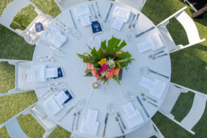 Outdoor Hotel Courtyard Boho Tropical Wedding Reception with Round White LInen Tables and Folding Chairs, Pink and Orange and Yellow Floral with Tropical Greenery Centerpiece, and Navy Blue and White Printed Coozie Wedding Favors| St Pete Beach Wedding Venue The Postcard Inn