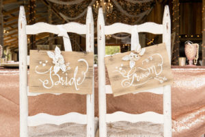 Rustic Wedding Reception Sweetheart Table with Vintage White Painted Ladderback Chairs, Hand-painted Bride and Groom Signs with White Ribbon and Roses, Sequin Rose Gold Table linen, and String Lights | Tampa Bay Venue Wishing Well Barn