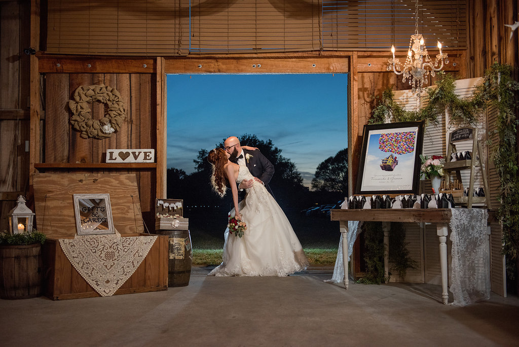 Bride and Groom Portrait at Barn Wedding Reception with Favor Table with Framed Disney Up Movie Poster, and Black and White Favors, with Vintage Chandelier, Wooden Table and dTrunk with Lace, Lantern, and Greenery Garland | Tampa Bay Venue Wishing Well Barn
