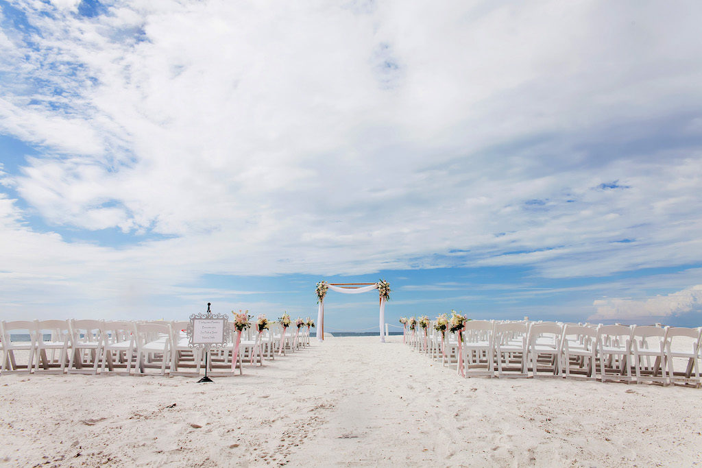 Outdoor Beach Wedding Ceremony decor with White Folding Chairs, White Floral with Pink Ribbon and Draping, Bamboo Ceremony Arch, and Framed Welcome Sign | Tampa Bay Waterfront Hotel Wedding Venue Hilton Clearwater Beach | Rentals A Chair Affair