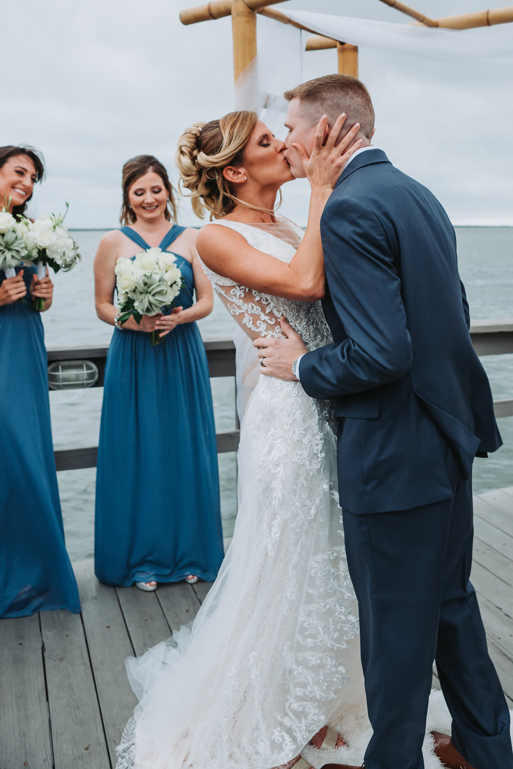 Outdoor Waterfront Wedding Ceremony First Kiss Portrait, Groom in Blue Suit, Bride in Lace Cutout Dress, Bridesmaids in Mismatched Blue Dresses with White and Succulent Greenery Bouquet | Tampa Bay Wedding Photographer Grind and Press Photography