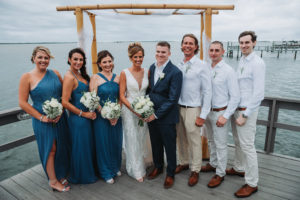 Outdoor Waterfront Wedding Party Portrait, Groom in Blue Suit with Boutonniere and Brown Shoes, Bride in Lace Cutout V Neck Dress, Bridesmaids in Mismatched Blue Dresses with White and Succulent Greenery Bouquet, Groomsmen in White Shirts with Khaki Pants | Tampa Bay Wedding Photographer Grind and Press Photography | Dunedin Venue Beso Del Sol