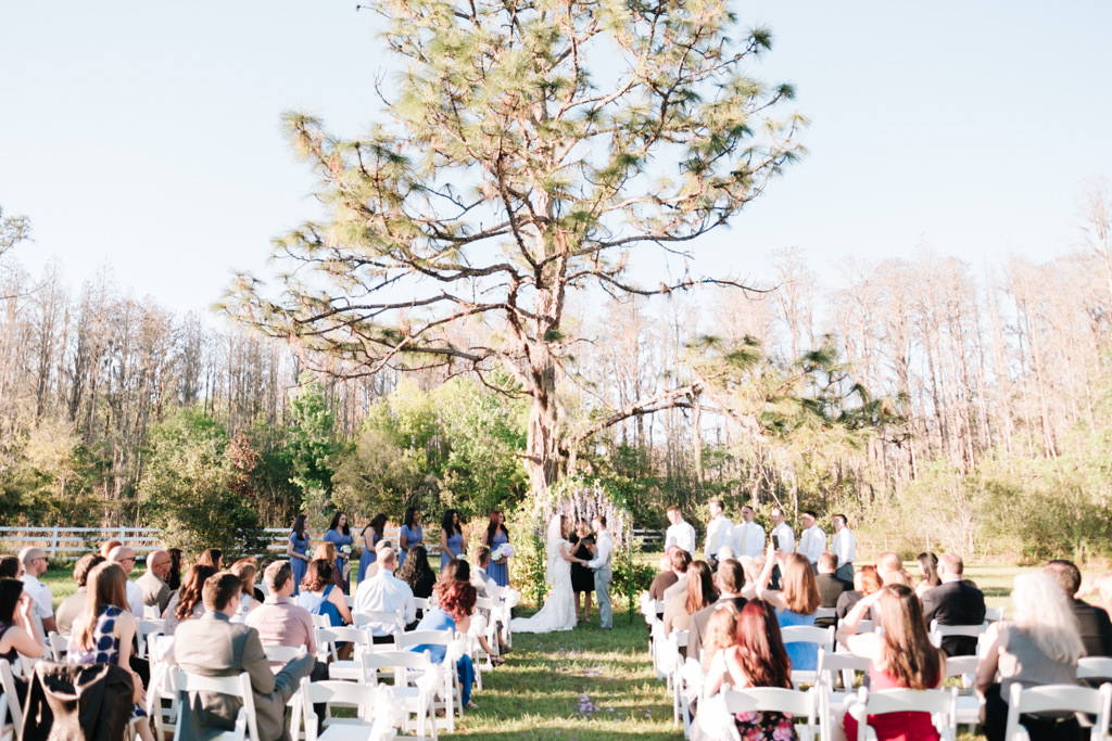 Outdoor Rustic Chic Wedding Ceremony under a Tree with White Folding Chairs, Bridesmaids in Lilac Blue Dresses, Groomsmen in Light Gray Suits | Tampa Bay Farm Wedding Venue Southern Plantation Oasis
