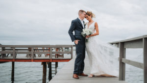 Outdoor Waterfront Wedding Portrait, Groom in Blue Suit with White Rose Boutonniere, Bride in Cutout V Neck Lace Dress with White Rose and Greenery Bouquet | Tampa Bay Wedding Photographer Grind and Press Photography | Dunedin Venue Beso Del Sol