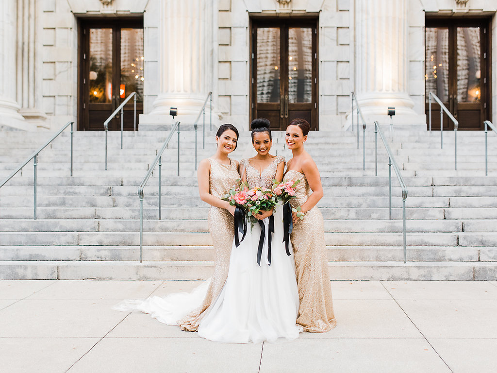 Outdoor Bridal Party Portrait, Bride with Bun Updo, Beaded Bodice Wedding Dress with Full Layered Skirt and Floral Lace Veil, Bridesmaids in Mismatched Column Gold Sequin Dresses, with Pink and Orange Floral Bouquet with Greenery and Long Black Ribbon | Downtown Tampa Historic Hotel Wedding Venue Le Meridien | Hair and Makeup Michele Renee The Studio