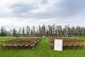 OUtdoor Farm Wedding Ceremony with Wooden Crossback Chairs, White Protea and Greenery Florals, and Wooden and White with Stylish Navy Blue Printed Welcome Sign, and Wooden Ceremony Arch with Marsala Red and Blush Pink Florals | Tampa Bay Wedding Planner NK Productions