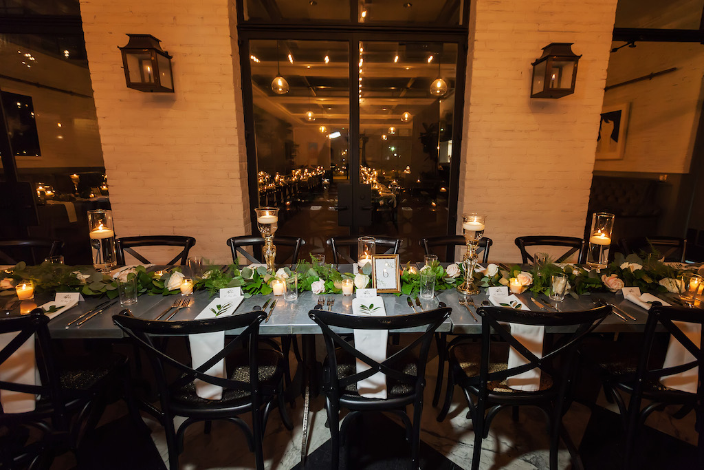Modern Industrial Chic Wedding Reception Feasting Table with Black Crossback Chairs, Low White Floral and Greenery Garland Centerpiece, White Linens, Floating Votive Candles in Clear Glass Candlestick Holders, Framed Table Number | Downtown Tampa Venue Oxford Exchange