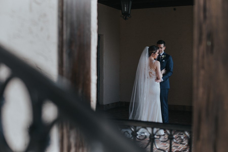 Outdoor Courtyard First Look Portrait, Bride with Long Comb Veil in Lace Applique Essence of Australia Dress, Groom in Navy Blue Suit with White Rose Boutonniere | Sarasota Wedding Photographer Brandi Image Photography