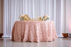 Elegant Romantic Wedding Reception Sweetheart Table with Rose Textured Pink LInen, Small White, Pink and Greenery Floral, White Draping, and Oversized Gold Bride and Groom Sing with Heart