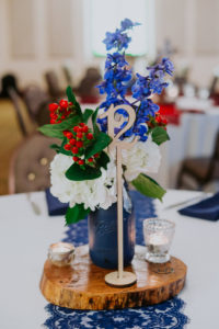 Americana Inspired Vintage Low Wedding Centerpiece with Blue and White Flowers and Red Berries with Greenery in Painted Vase on Natural Wood Round, with White Linens and Blue Lace Runner and Laser Cut Wooden Table Number | Tampa Bay Wedding Planner Special Moments Event Planning | St Pete Florist Apple Blossoms Floral Designs
