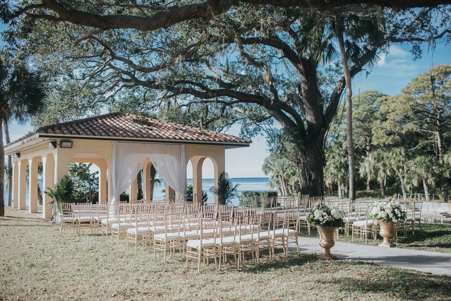 Outdoor Southern Waterfront Wedding Ceremony with Gold Chiavari Chairs, Large Classical Planters with White FLoral and Greenery, and White Draping Ceremony Arch in Gazebo | Bradenton Wedding Venue Powel Crosley Estate